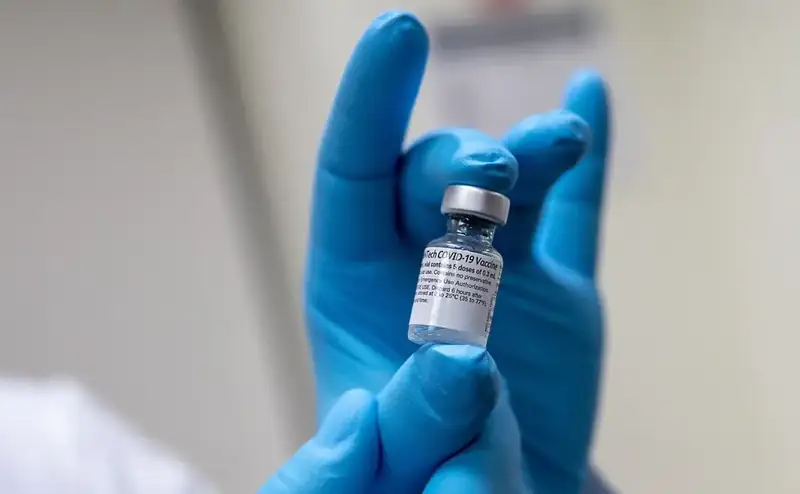 Een hand in een blauwe handschoen houdt een flacon coronavaccin vast