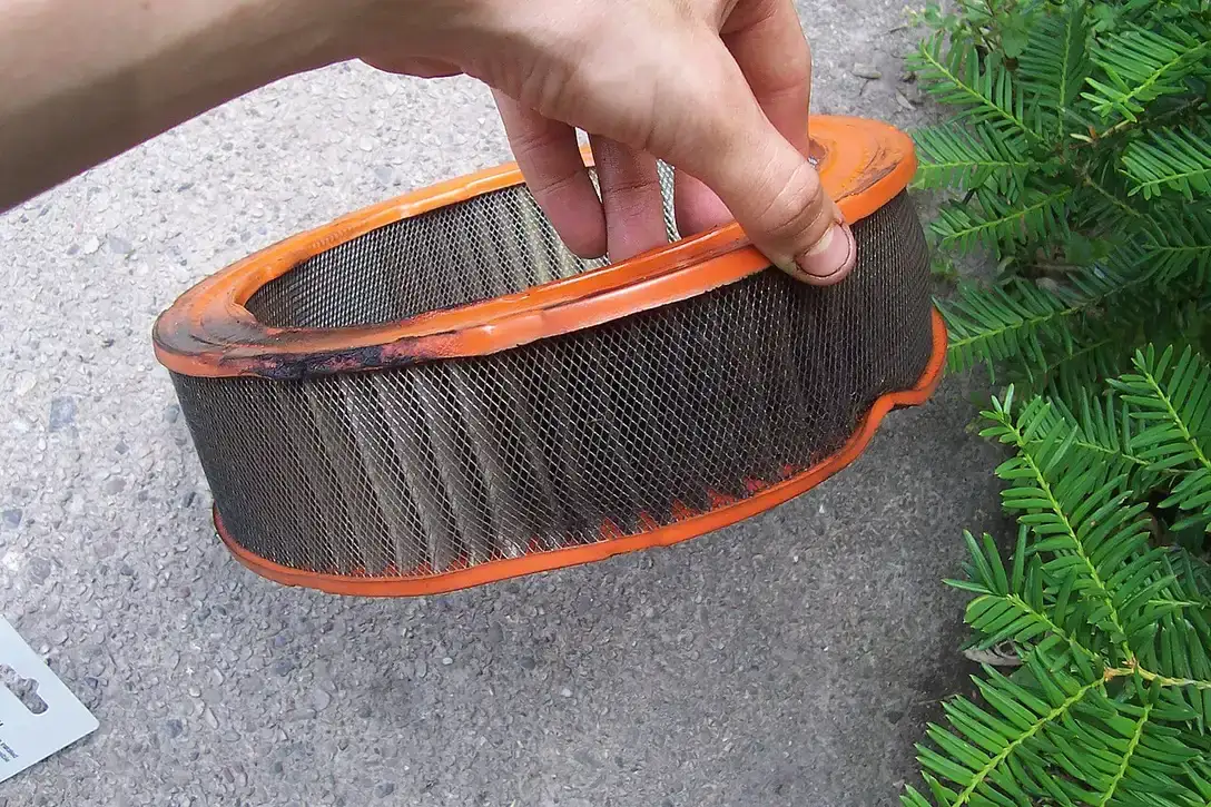 Hand houdt een rond, oranje luchtfilter vast waarvan het filtermateriaal grijs en zwart is van het vuil