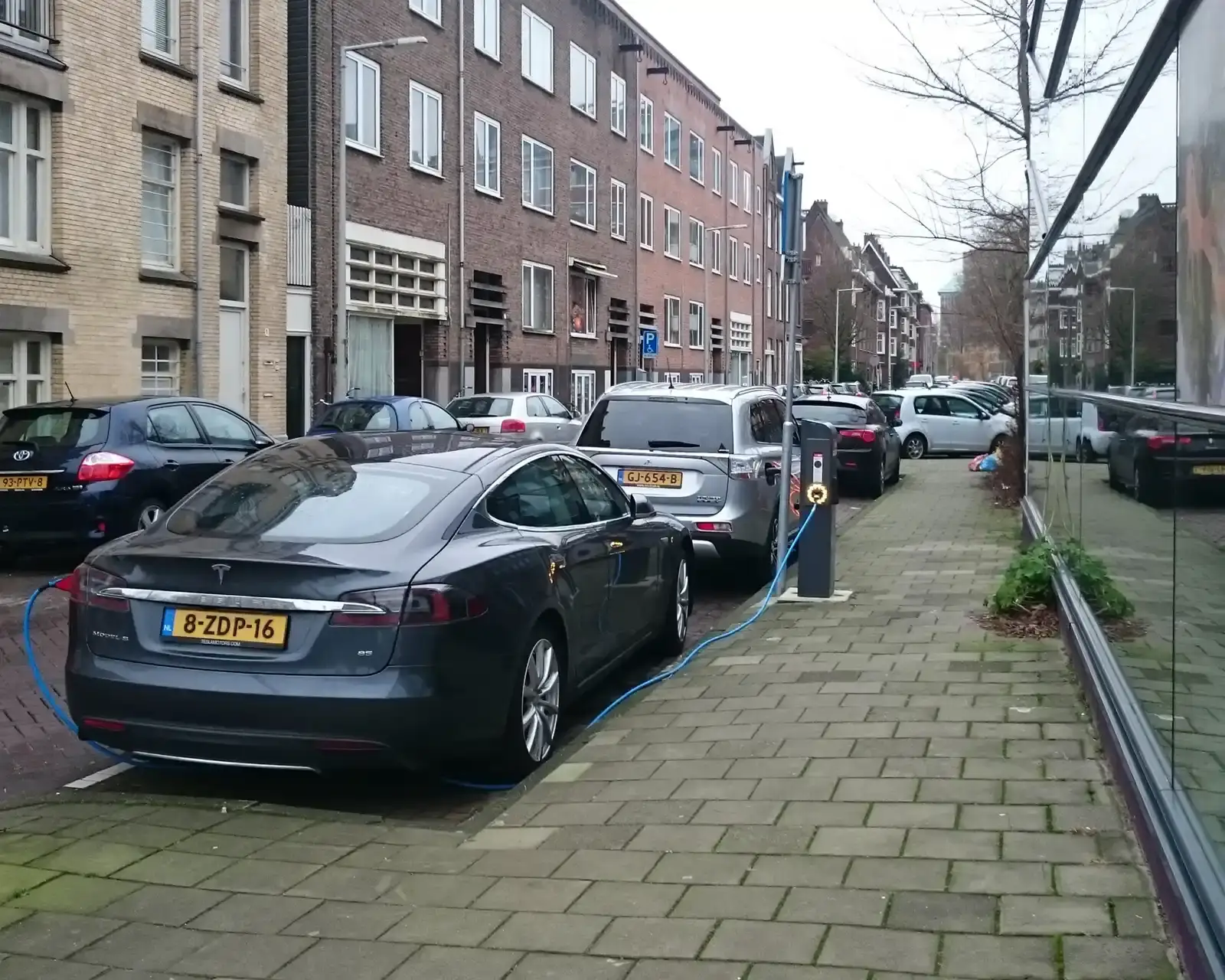 Een elektrische auto en een plug-in hybride staan aan een openbare laadpaal in een straat in Amsterdam