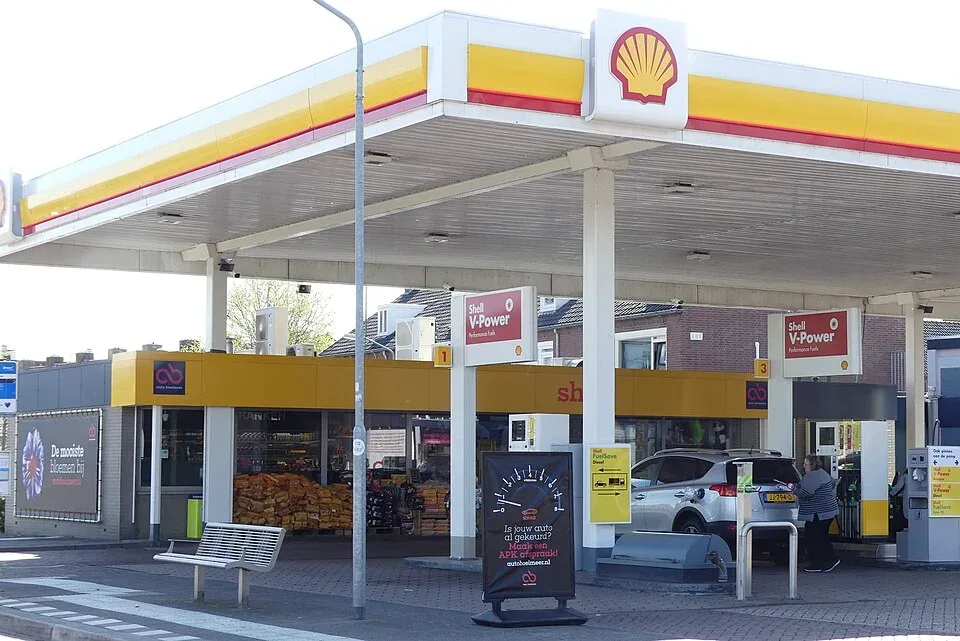 Shell-tankstation in een Nederlandse woonwijk met geel-rode overkapping, pompen en een vrouw die een zilveren auto tankt